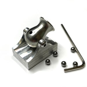 Op de afbeelding: Een klein, zilveren metalen kanon gemonteerd op een basis met een zilveren metalen inbussleutel en verschillende zilveren metalen ballen.