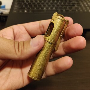 May include: A gold-colored metal lighter with a textured finish and a small window on the side. The lighter is held in a hand.
