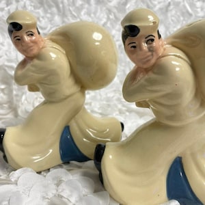 Könnte beinhalten: Zwei Keramikfiguren von Männern, die Säcke tragen. Die Figuren tragen cremefarbene Uniformen mit blauen Akzenten und schwarzen Schuhen. Die Gesichter sind detailliert bemalt und sie tragen passende Mützen. Die Figuren stehen auf einer weißen, strukturierten Oberfläche.