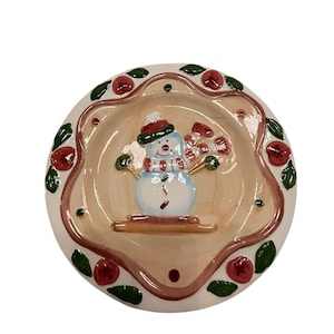 May include: A round, ceramic Christmas dish featuring a snowman design. The snowman has a red hat and scarf, with a light blue body. The rim is decorated with red berries and green leaves. The dish has a beige interior.