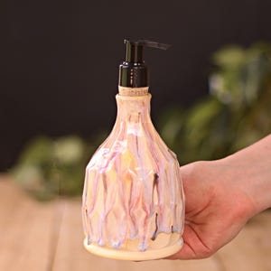 May include: A ceramic soap dispenser with a black pump. The dispenser has a cream base and is decorated with dripped glaze in pink, purple, and yellow. The dispenser is held by a hand against a blurred background of green and brown.