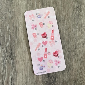 May include: A pink card featuring illustrations of beauty products and gifts, such as lipstick, bows, and hearts. The card has the text "TheCashCubbie" at the bottom. The card is on a wooden surface.