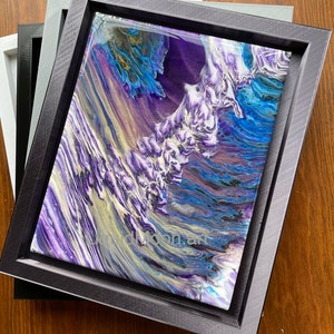 Op de afbeelding: Abstracte kunstprint in een zwarte lijst. Het kunstwerk heeft een golfachtig ontwerp met tinten paars, blauw en wit. De tekst "dmoon art" is zichtbaar in de linker benedenhoek. De lijst is op andere lijsten gestapeld.