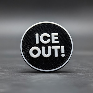 May include: A circular button with the text "ICE OUT!" in white against a black background. The button has a white border and a textured surface. The background is a plain grey.