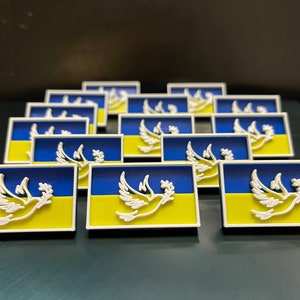 Puede incluir: Un grupo de 18 pequeñas chapas rectangulares impresas en 3D que muestran la bandera ucraniana con una paloma blanca de la paz en el centro. Las chapas están dispuestas en un patrón escalonado.