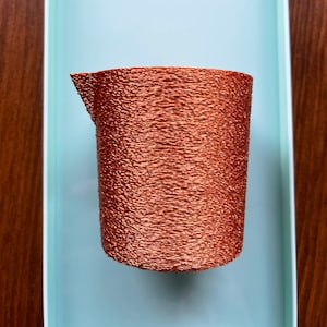 Pode incluir: Um recipiente cilíndrico de cor cobre, texturizado, com uma borda superior ligeiramente irregular.