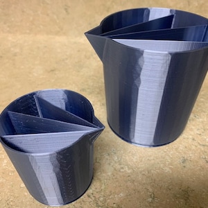 May include: Two dark blue and grey paint pouring cups. The cups are made of plastic and have a unique design with internal dividers. The cups are designed to help with paint pouring techniques.