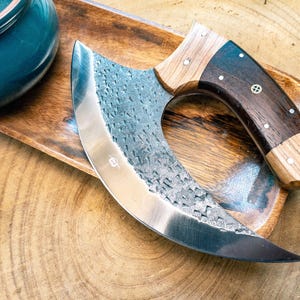 May include: A curved, stainless steel knife with a dark wood handle and a light wood bolster. The blade has a textured surface. The knife rests on a wooden tray, with a teal ceramic bowl in the background. The knife is designed for chopping.