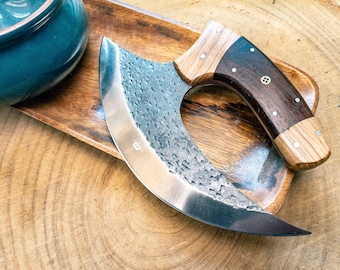 May include: A curved, stainless steel knife with a dark wood handle and a light wood bolster. The blade has a textured surface. The knife rests on a wooden tray, with a teal ceramic bowl in the background. The knife is designed for chopping.