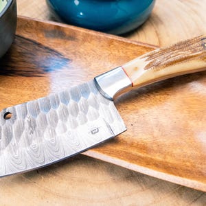 May include: A cleaver knife with a patterned blade and a light-colored handle with a dark brown pattern. The knife has a silver bolster and a small hole near the blade's edge. It rests on a wooden tray.