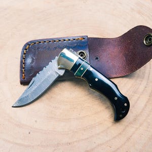 May include: A black and gold pocket knife with a damascus steel blade and a leather sheath.
