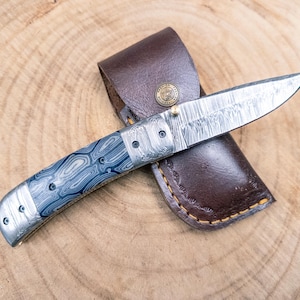May include: A folding knife with a patterned steel blade and a blue and silver handle, resting on a brown leather sheath. The knife has a decorative design and is suitable for outdoor use.