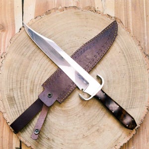 May include: A silver knife with a brown leather sheath, lying on a wooden surface. The knife has a wooden handle with a dark brown finish.