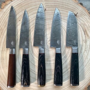 May include: Five kitchen knives with black and brown wooden handles on a wooden surface. The blades are made of stainless steel and have a hammered finish.