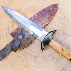 May include: A handmade dagger with a polished steel blade and a wooden handle. The handle has black accents and brass rivets. The dagger rests on a brown leather sheath with a belt loop. The sheath is decorated with a stitched pattern.
