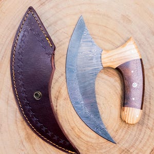 May include: A curved, Damascus steel knife with a wooden handle and a brown leather sheath. The blade has a distinctive pattern, and the handle features a combination of light and dark wood. The sheath is stitched with a decorative pattern.