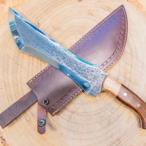 May include: A handcrafted knife with a blue-toned blade and a wooden handle, resting on a brown leather sheath. The knife is displayed on a wooden surface, showcasing its intricate design and craftsmanship. The blade measures approximately 12 inches.
