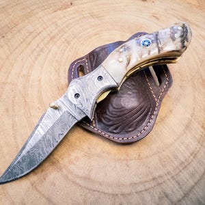May include: A Damascus steel folding knife with a bone handle and a blue gem inlay. The knife rests on a brown leather sheath with decorative tooling. The blade is open, showcasing its intricate pattern.