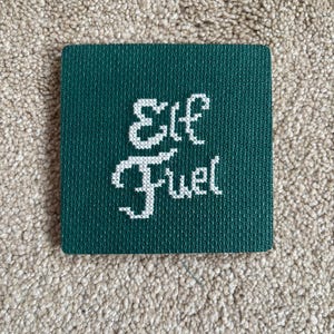 May include: A square, dark green coaster with the words "Elf Fuel" stitched in white. The coaster is made of a textured material and is displayed on a neutral-colored, textured surface.