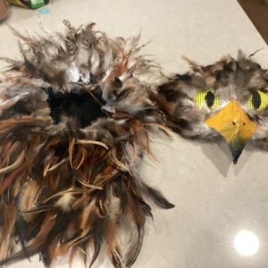 Könnte beinhalten: Eine handgefertigte Vogelmaske mit braunen, beigen und schwarzen Federn. Die Maske hat einen gelben Schnabel, gelbe Netzk Augen und einen gefiederten Hals. Dieses Kostümzubehör ist ideal für Halloween oder Theateraufführungen.