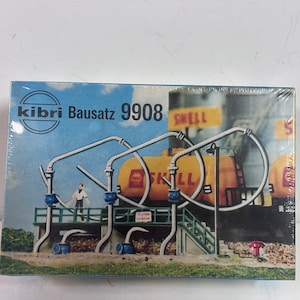 May include: A model kit box labeled "kibri Bausatz 9908" featuring a miniature Shell oil refinery scene. The scene includes yellow oil tanks, silver pipes, a small figure, and a green platform. The box is sealed in clear plastic.