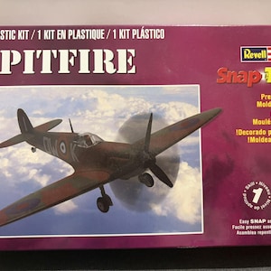 May include: A Revell Snap Tite model kit of a Spitfire airplane. The box is primarily burgundy with the plane's image in the center. The plane is brown and green with a propeller and wings. Text on the box includes "SPITFIRE" and "Pre-decorated!"