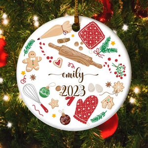 May include: A white ceramic Christmas ornament with baking-themed illustrations. It features a rolling pin, gingerbread man, and the name "emily" with the year "2023". The ornament is round and has a gold string for hanging.