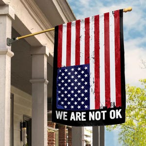May include: A distressed American flag with a black banner at the bottom that reads "WE ARE NOT OK". The flag is hanging from a pole outside a house.