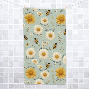 May include: A light blue towel with a floral pattern of yellow sunflowers, white daisies, and cartoon bees. The words "fuck trump" are written throughout the design. The towel is hanging on a clothesline.