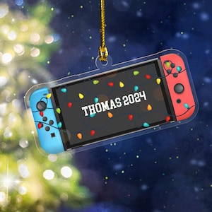 May include: A clear acrylic Christmas ornament shaped like a video game console. The console has blue and red controllers, with the text "THOMAS 2024" on the screen. Colorful string lights are draped across the screen. A gold string is attached for hanging.