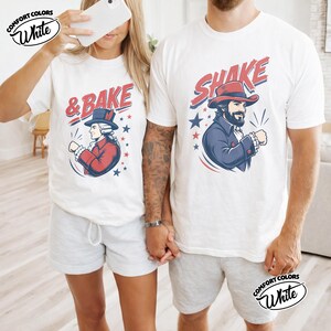 May include: Two white t-shirts featuring graphic prints. The left shirt displays a historical figure in a red coat and top hat with the text & BAKE. The right shirt shows a bearded figure in a blue coat and red hat with the text SHAKE.