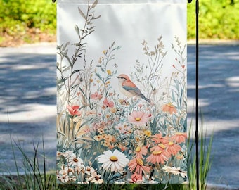 May include: A garden flag with a white background and a colorful floral design. A small blue bird is perched on a branch in the center of the flag. The flowers are in shades of pink, purple, and yellow.