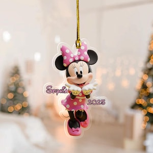 May include: A Minnie Mouse ornament with a pink polka dot bow and dress. The ornament has the name "Sophia" and the year "2025" printed on it. It is hanging from a gold string.