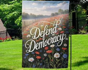 May include: A garden flag with the words "Defend Democracy" in white script. The flag features a field of colorful wildflowers in red, white, and blue, with a scenic background of a fence and trees under a cloudy sky.