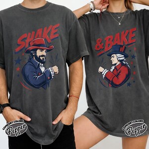 May include: Two charcoal gray t-shirts featuring a graphic print. The left shirt displays a man in a red hat with the text SHAKE. The right shirt shows a man in a blue hat with the text & BAKE. Both shirts include red and blue star accents.