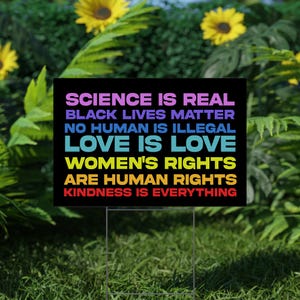 May include: A black and white yard sign with rainbow colored text that reads "Science is real, Black Lives Matter, No Human is Illegal, Love is Love, Women's Rights are Human Rights, Kindness is Everything."