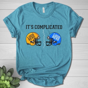 May include: A turquoise t-shirt with the text "IT'S COMPLICATED" above two football helmets. One helmet is yellow with the text "LOGO HERE", and the other is blue with the text "LOGO HERE". A white zig-zag line separates the helmets.