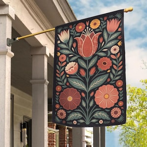 May include: A decorative garden flag featuring a floral design with a large central flower and various smaller flowers in shades of red, orange, and white against a dark background. The flag is hanging from a gold-colored pole.