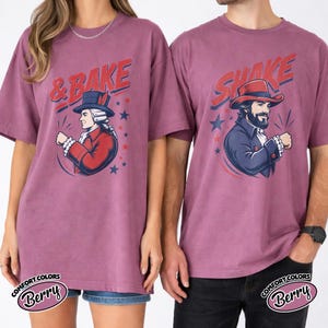 May include: Two berry-colored t-shirts featuring graphic prints. The left shirt displays a person in a colonial-style red coat and hat with the text & BAKE. The right shirt shows a person in a blue coat and hat with the text SHAKE.