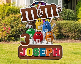 May include: A yard sign featuring the M&M's logo and characters, with the number 3 and the name JOSEPH. The sign has a brown border and a green lawn background. The M&M's characters are in various colors.