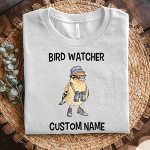 May include: A white t-shirt with a cartoon bird wearing a hat and glasses and holding binoculars. The text "Bird Watcher" and "Custom Name" is printed on the shirt.