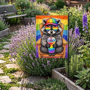 May include: A rectangular garden flag featuring a gray raccoon wearing a rainbow-colored baseball cap and heart-shaped sunglasses. The raccoon sits in a wooden chair, holding a rainbow-colored drink with a straw against a vibrant rainbow background.