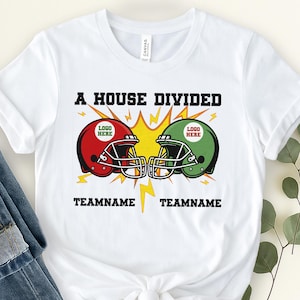 May include: White t-shirt featuring a graphic with two football helmets, one red and one green, facing each other. The text "A HOUSE DIVIDED" is above the helmets, with "TEAMNAME" below each helmet. The design includes lightning bolts and a yellow burst.