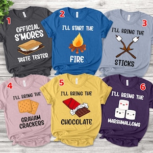 May include: Six different t-shirts with text and graphics related to making s'mores. The shirts are in various colors, including gray, blue, light pink, yellow, purple, and light blue. The text on the shirts includes "Official S'mores Taste Tester", "I'll Start the Fire", "I'll Bring the Sticks", "I'll Bring the Graham Crackers", "I'll Bring the Chocolate", and "I'll Bring the Marshmallows".