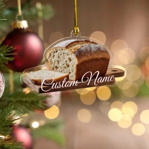May include: A clear acrylic Christmas ornament shaped like a loaf of bread and a slice on a wooden cutting board. The ornament has a gold string for hanging and the words "Custom Name" in a script font.
