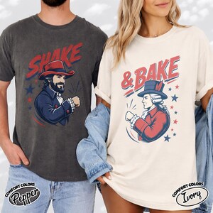 May include: A charcoal gray t-shirt with the red text SHAKE and a graphic of a man in a colonial hat, and an ivory t-shirt with the red text & BAKE and a graphic of a colonial figure in a hat, both featuring blue stars and red accents.
