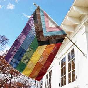 May include: A rectangular flag featuring a patchwork design of various fabric textures and patterns in a rainbow color spectrum. The flag includes a triangular chevron section on the left side with brown, black, white, pink, and light blue fabric patches.
