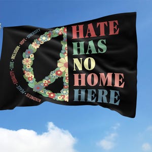 May include: Black flag with the words "HATE HAS NO HOME HERE" in colorful letters. A peace symbol made of flowers is on the left. The flag also has words like "Kindness, Peace, Equality, Love, Inclusion, Hope, Diversity."