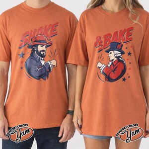 May include: Two burnt orange t-shirts featuring graphic prints. The left shirt displays a bearded figure in a blue colonial coat with the text SHAKE. The right shirt shows a figure in a red colonial coat with the text & BAKE. Both include stars.