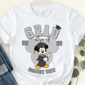 May include: White t-shirt featuring a graduation design with the text "GRAD class of 26" in gray block letters. Mickey Mouse in a graduation gown and cap is in the center. The words "CUSTOM TEXT" are at the bottom.